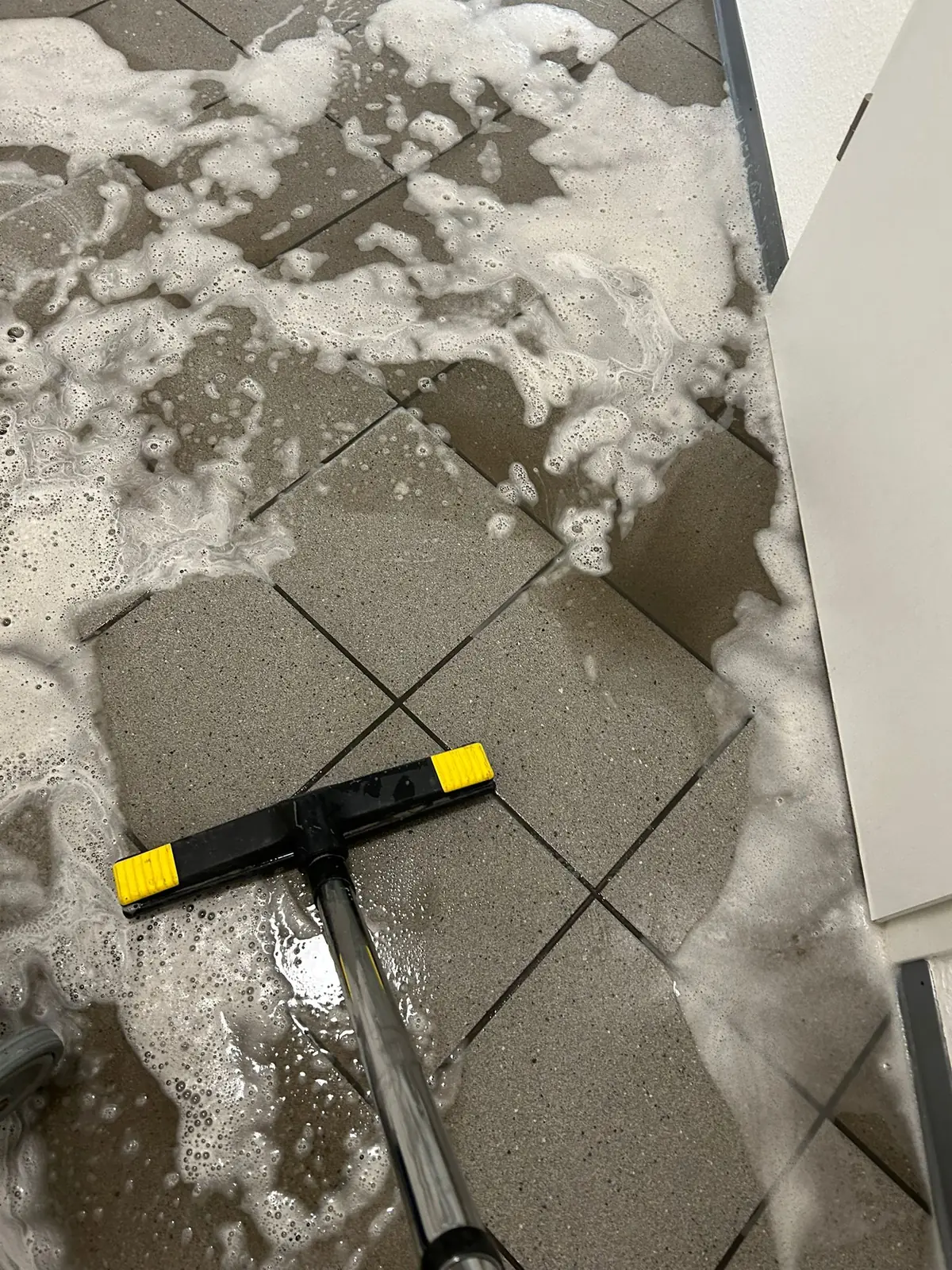 Mit einem schwarz-gelben Wasserschieber am Stiel wird Reinigungsschaum über einen grauen Fliesenboden zusammengezogen